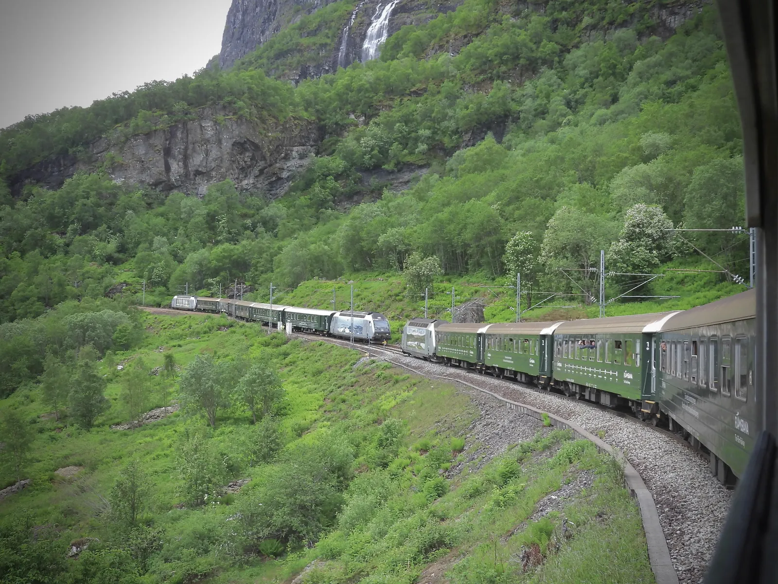 Two dark green Flåm Railway trains passing each other on a curve of track through a steep green valley, with a waterfall on the cliff above