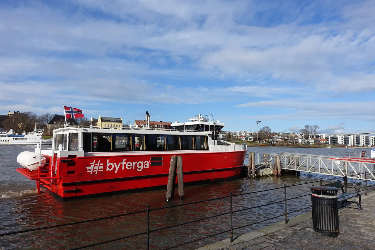A red and white city ferry marked 'byferga', flying a Norwegian flag, moored at a stone quay on the Glomma, with older houses and modern apartment blocks along the far bank