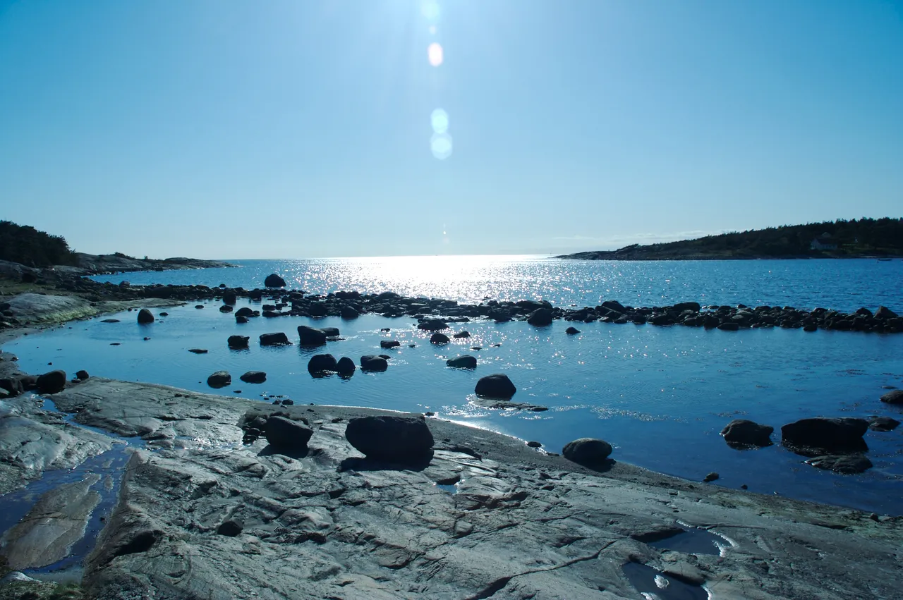 Smooth, bare granite rock and scattered boulders at the water's edge in Ytre Hvaler, with a calm, sunlit sea opening out between two low wooded islands