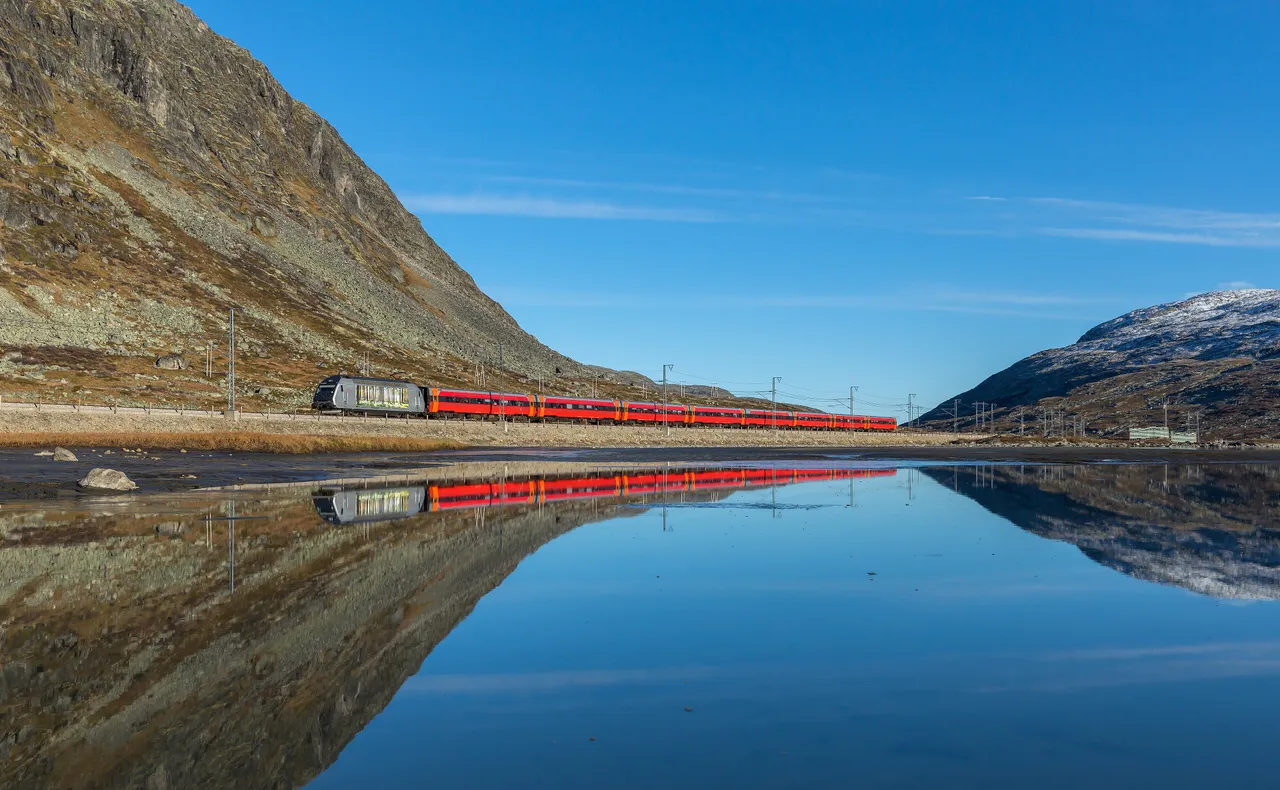 A red NSB passenger train hauled by a grey locomotive running along the shore of a still lake on a treeless mountain plateau, the train and the rocky slopes reflected in the water under a clear blue sky