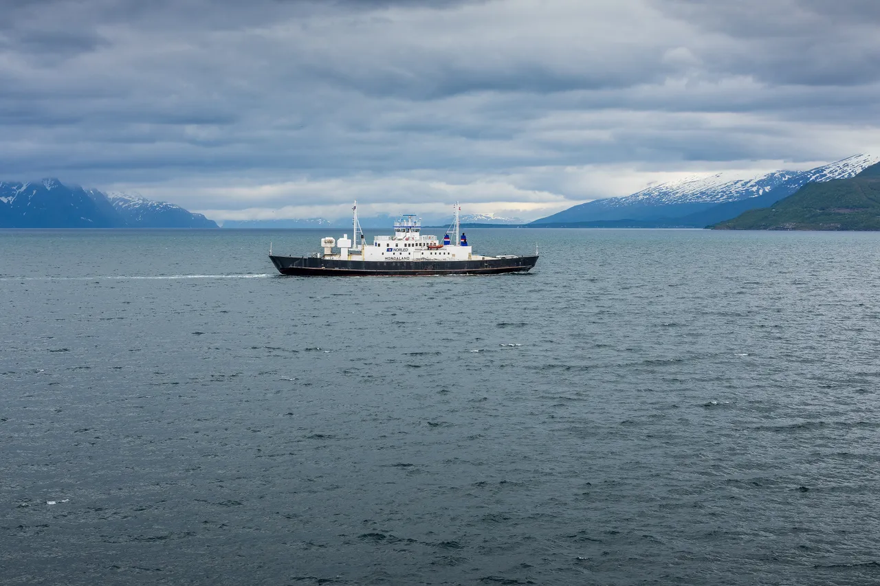 A small black-and-white car ferry with the Norled logo crossing a wide, choppy fjord under a heavy grey sky, with snow-streaked mountains on both shores