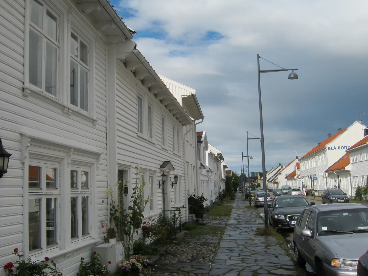 A cobbled pavement running past a row of white-painted wooden houses with flowers by the doors, on a quiet street in Posebyen under a cloudy sky