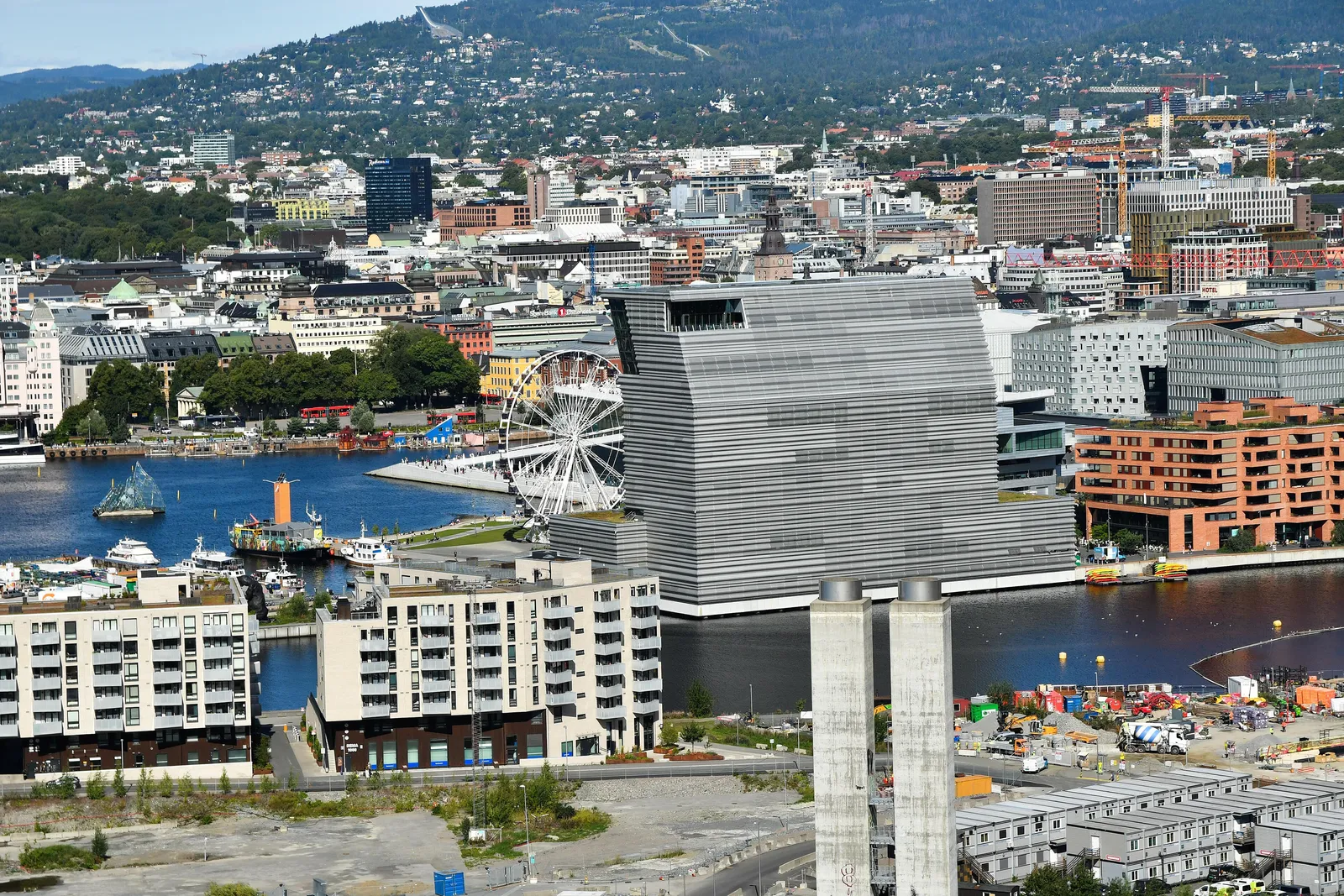 Central Oslo seen from the Ekeberg hillside on a summer day, with the tall grey MUNCH museum on the harbour in Bjørvika, the white Opera House roof and a Ferris wheel beside it, the city centre behind and the Holmenkollen ski jump on the wooded ridge above