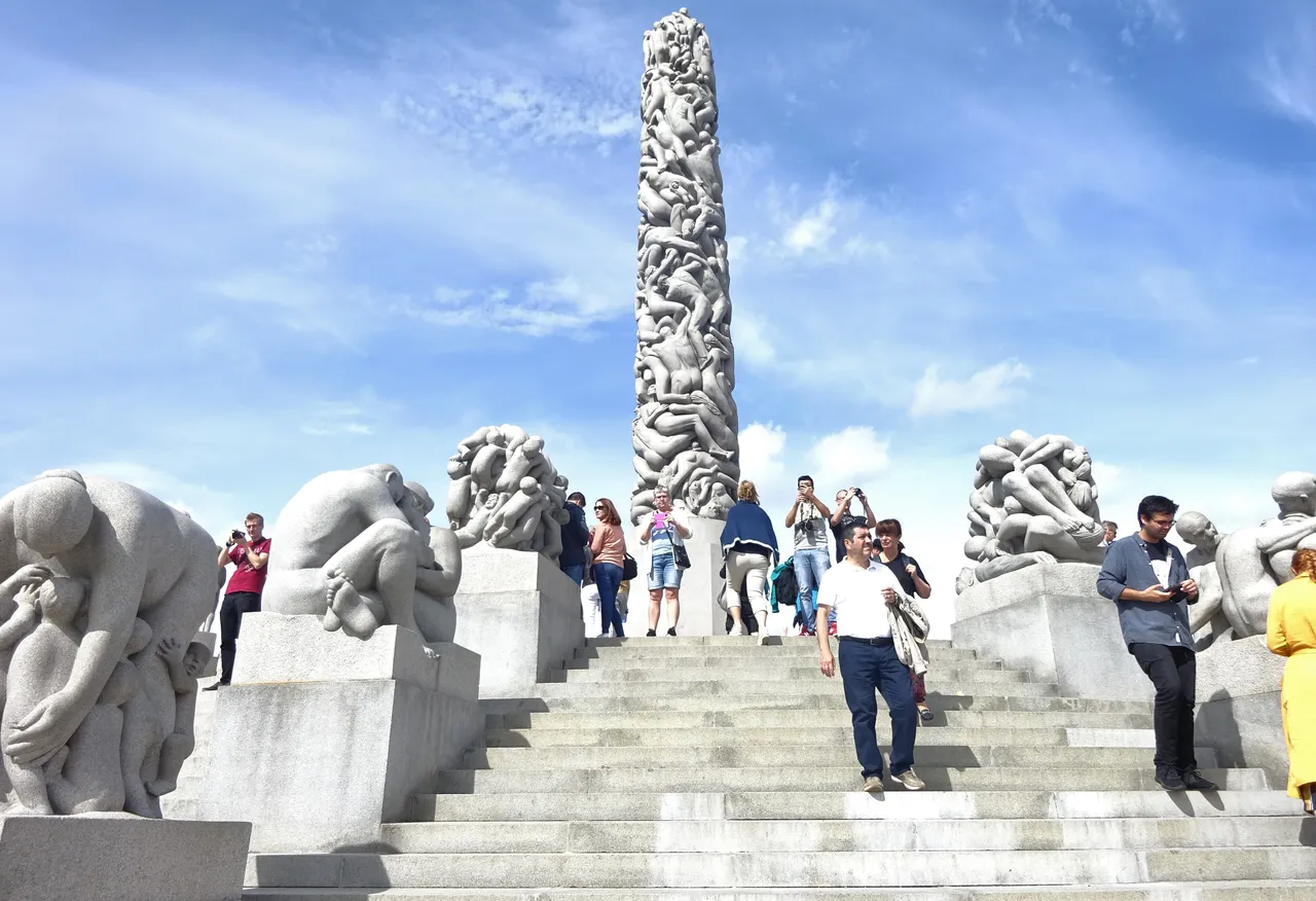 Visitors on the granite steps leading up to the Monolith in Vigeland Park, a tall column of carved human figures, flanked by granite sculpture groups under a blue sky