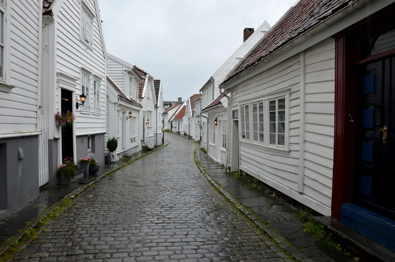 A narrow, wet cobbled lane between white-painted wooden houses with red tile roofs, lanterns and pots of pink flowers by the doors