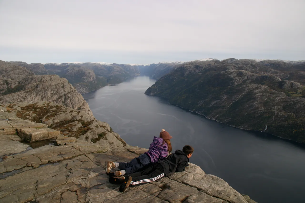 Two hikers lying on bare grey rock at the edge of a cliff, looking down a long, dark fjord between steep rocky hills with a dusting of snow on the tops