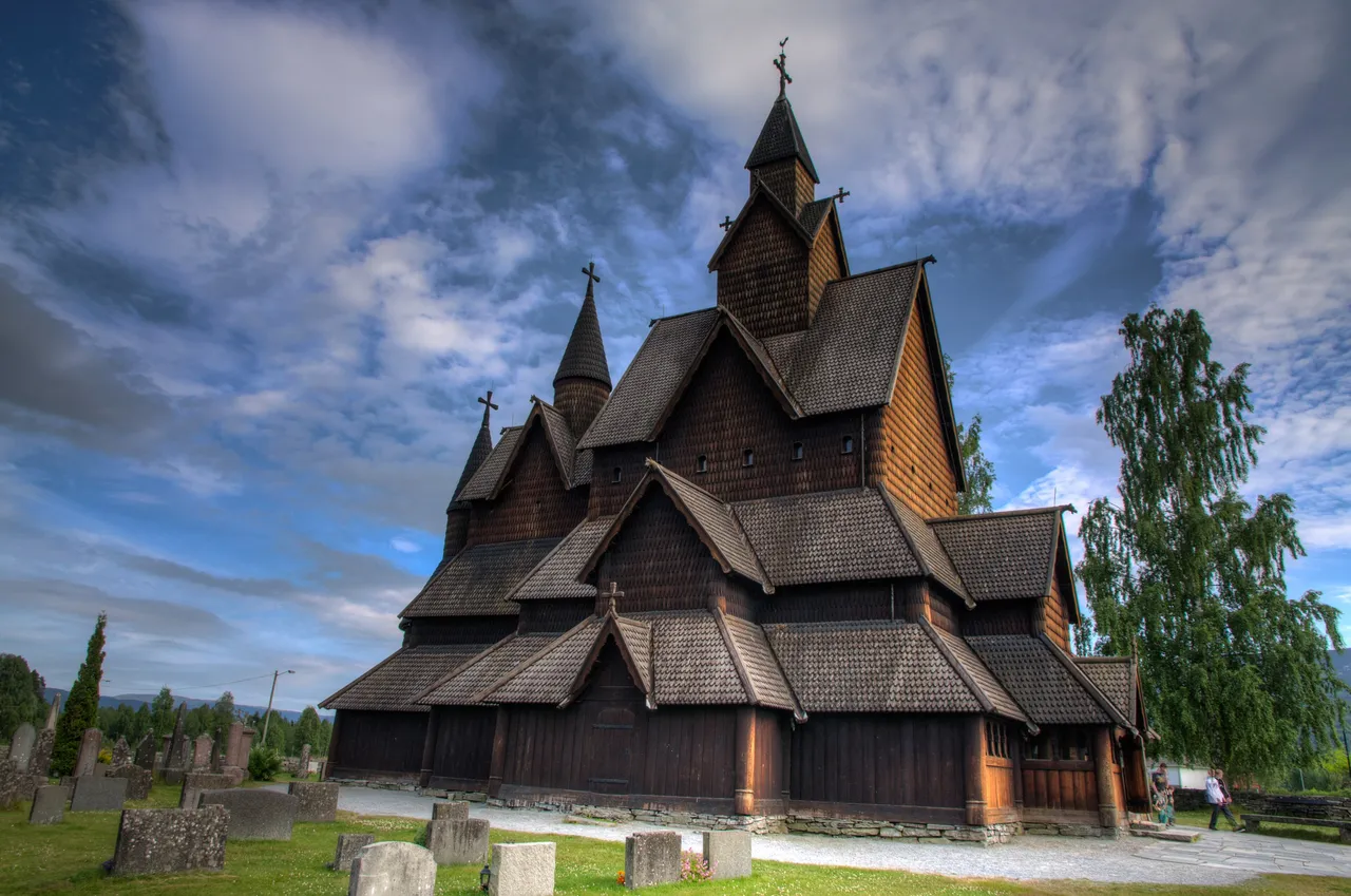 Heddal Stave Church under a blue sky with white clouds, a large dark-brown shingled church with many gables, three spires and a gallery around the base, gravestones in front