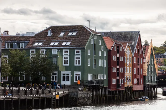 Old wooden warehouses painted green, dark red and ochre standing on wooden piles along the Nidelva river in Trondheim, with people sitting on a waterside boardwalk under a cloudy sky